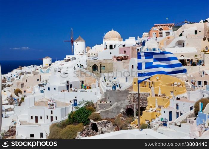 Oia town on Santorini island, Greece. Famous windmills on cliff over the Caldera, Aegean sea. Focus on waving Greek flag