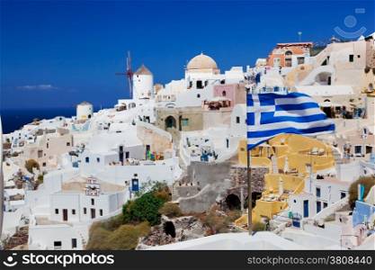 Oia town on Santorini island, Greece. Famous windmills on cliff over the Caldera, Aegean sea. Focus on waving Greek flag