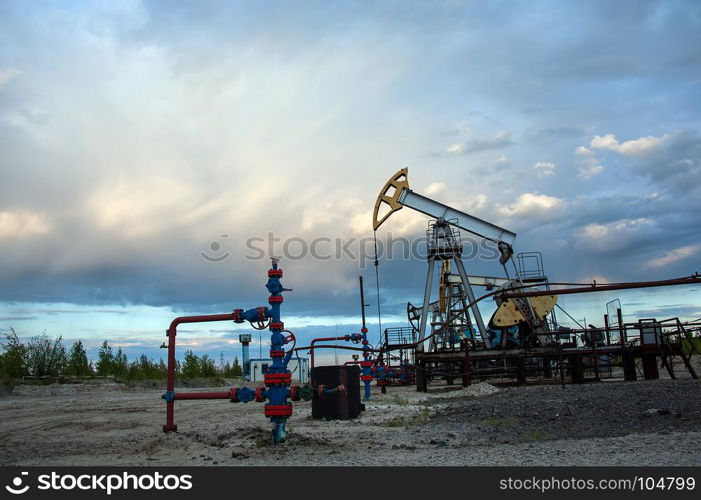 Oil pump jack and wellhead with valve armature during sunset on the oilfield. Extraction of oil. Oil and gas concept. Dramatic cloudy sky background.