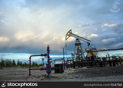 Oil pump jack and wellhead with valve armature during sunset on the oilfield. Extraction of oil. Oil and gas concept. Dramatic cloudy sky background.