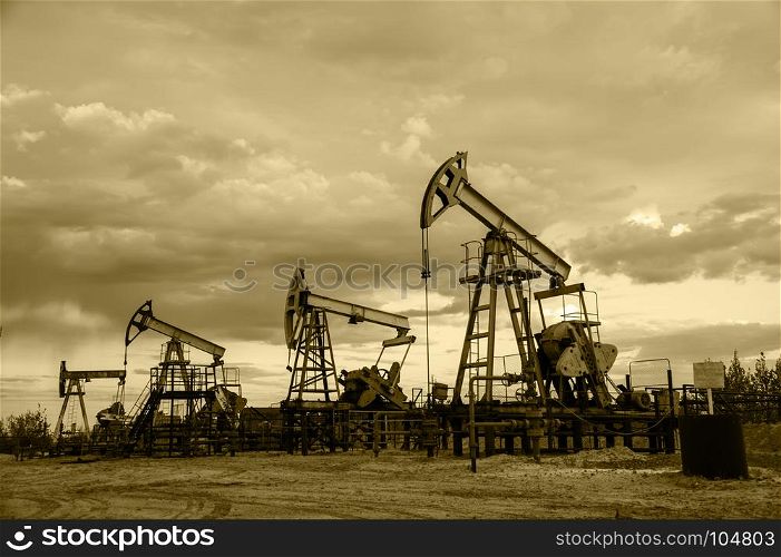 Oil pump jacks group and wellhead with valve armature during sunset on the oilfield. Oil and gas concept. Dramatic cloudy sky background. Toned sepia.