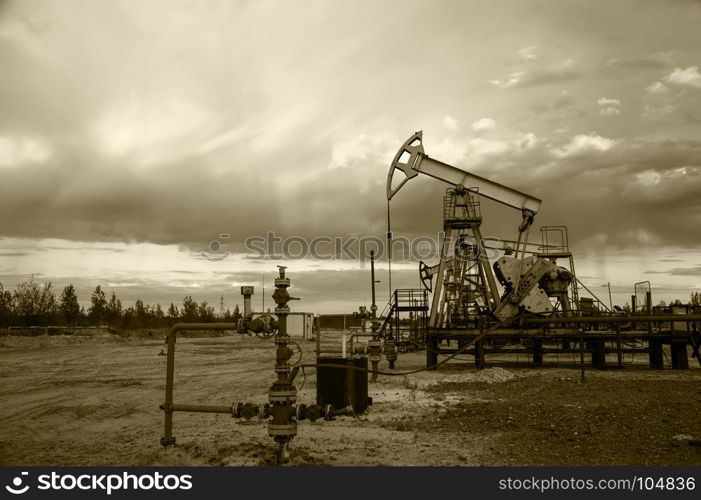 Oil pump jacks group and wellhead with valve armature during sunset on the oilfield. Oil and gas concept. Dramatic cloudy sky background. Toned sepia.