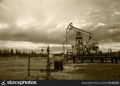 Oil pump jacks group and wellhead with valve armature during sunset on the oilfield. Oil and gas concept. Dramatic cloudy sky background. Toned sepia.