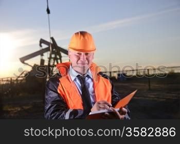Oil worker in orange uniform and helmet on of background the pump jack and sunset sky. Look into the camera.