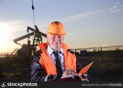 Oil worker in orange uniform and helmet on of background the pump jack and sunset sky. Look into the camera.