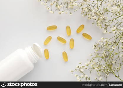Oil yellow gelatin capsules with white bottle on white background with flowers, vitamins and antioxidant concept Omega 3, liver cod or evening primose oil for healthcare. Minimalism. Copy space. Oil yellow gelatin capsules with white bottle on white background with flowers, vitamins and antioxidant concept Omega 3, liver cod or evening primose oil for healthcare. Minimalism. Copy space.