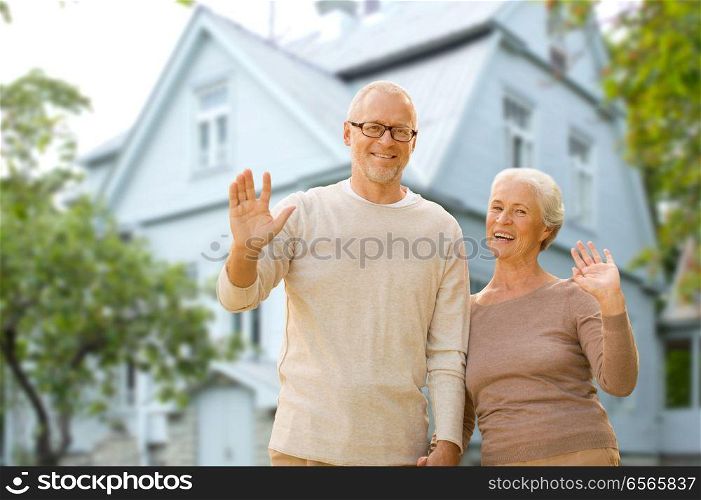 old age, accommodation and real estate concept - happy senior couple waving hands over living house background. happy senior couple waving hands over living house