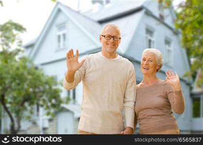 old age, accommodation and real estate concept - happy senior couple waving hands over living house background. happy senior couple waving hands over living house