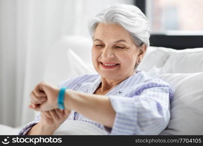 old age and people concept - happy smiling senior woman in pajamas with health tracker sitting in bed at home bedroom. happy old woman with health tracker sitting in bed