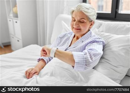 old age and people concept - happy smiling senior woman in pajamas with smart watch sitting in bed at home bedroom. happy senior woman sitting in bed at home bedroom