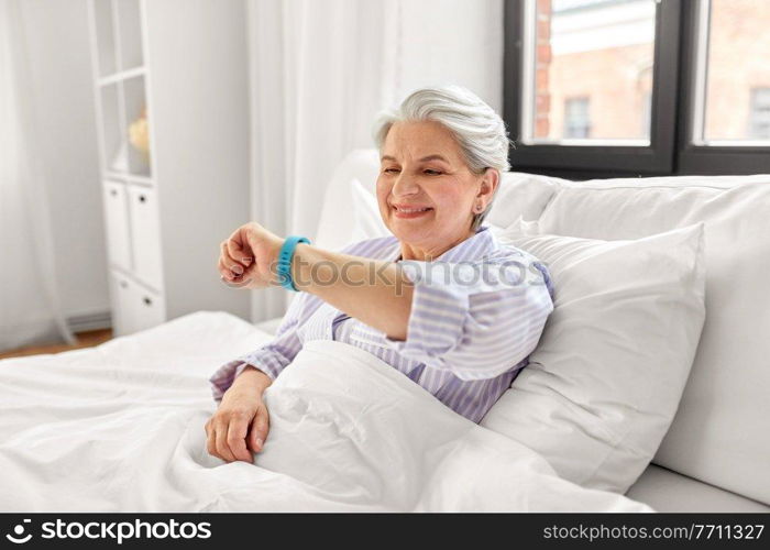 old age and people concept - happy smiling senior woman in pajamas with health tracker sitting in bed at home bedroom. happy old woman with health tracker sitting in bed