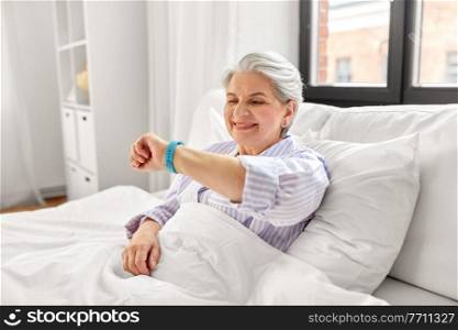 old age and people concept - happy smiling senior woman in pajamas with health tracker sitting in bed at home bedroom. happy old woman with health tracker sitting in bed