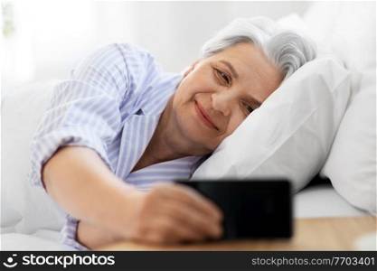 old age and people concept - happy smiling senior woman with smartphone lying in bed at home bedroom. happy senior woman with smartphone in bed at home