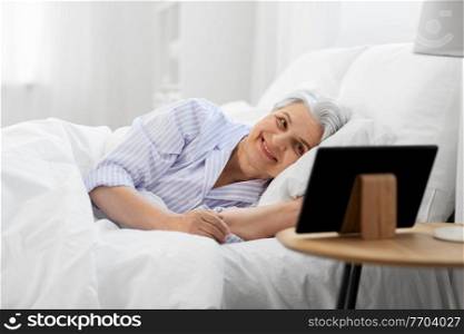 old age and people concept - happy smiling senior woman with tablet pc lying in bed at home bedroom. happy senior woman with tablet pc in bed at home