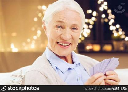 old age, gamble, holidays and people concept - happy smiling senior woman playing cards at home over garland lights background. happy senior woman playing cards on christmas