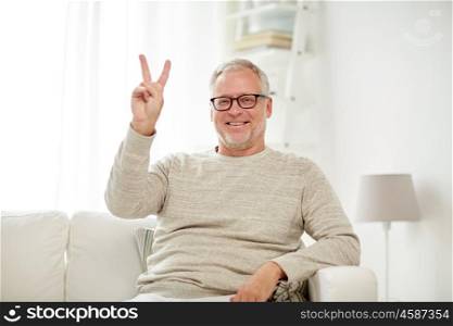 old age, gesture and people concept - smiling senior man in glasses sitting on sofa and showing v sign at home