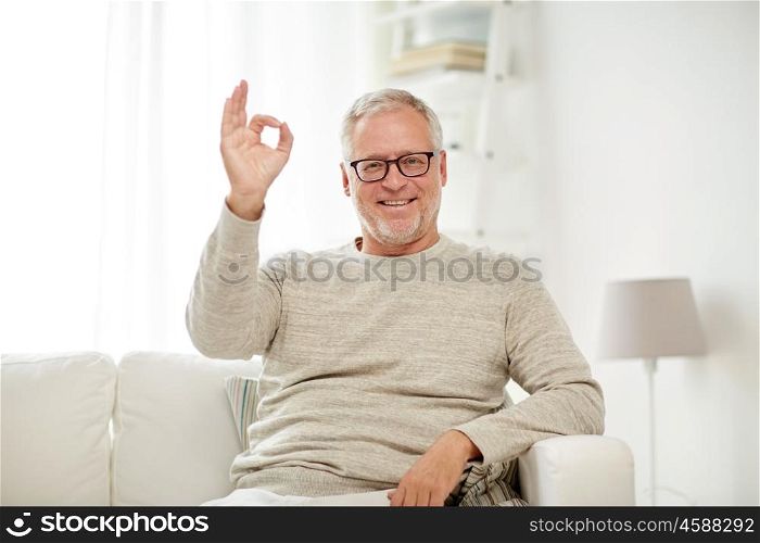 old age, gesture, comfort and people concept - smiling senior man in glasses sitting on sofa and showing ok hand sign at home