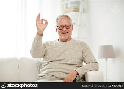 old age, gesture, comfort and people concept - smiling senior man in glasses sitting on sofa and showing ok hand sign at home