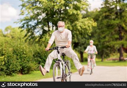 old age, health and pandemic concept - happy senior couple wearing face protective medical mask for protection from virus disease riding bicycles at summer park. senior couple in masks riding bicycles at park