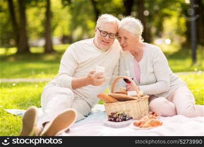 old age, holidays, leisure and people concept - happy senior couple with smartphone, picnic basket and wine sitting on blanket at summer park. senior couple with smartphone at picnic in park