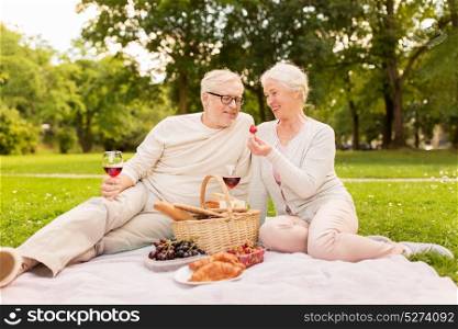 old age, holidays, leisure and people concept - happy senior couple with picnic basket and wine glasses sitting on blanket at summer park. happy senior couple having picnic at summer park