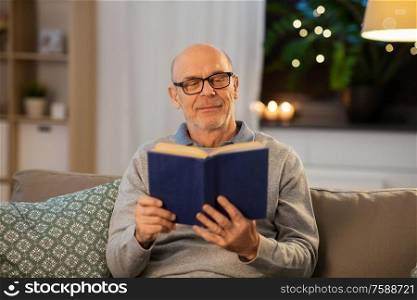 old age, leisure and people concept - happy bald senior man sitting on sofa and reading book at home in evening. happy bald senior man on sofa reading book at home