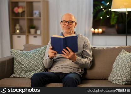 old age, leisure and people concept - happy bald senior man sitting on sofa and reading book at home in evening. happy bald senior man on sofa reading book at home