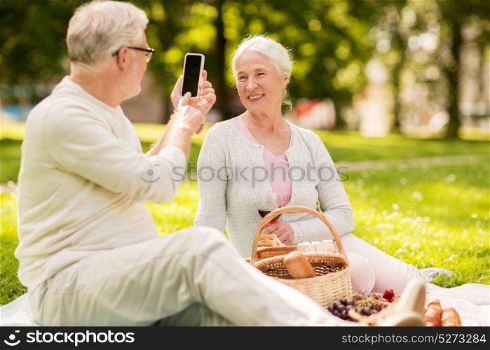 old age, leisure and people concept - happy senior couple with picnic basket and wine taking picture by smartphone at summer park. senior couple taking picture by smartphone at park