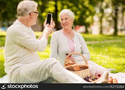 old age, leisure and people concept - happy senior couple with picnic basket and wine taking picture by smartphone at summer park. senior couple taking picture by smartphone at park