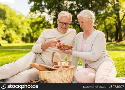 old age, leisure and people concept - happy senior couple with picnic basket eating strawberries at summer park. senior couple with strawberries at picnic in park
