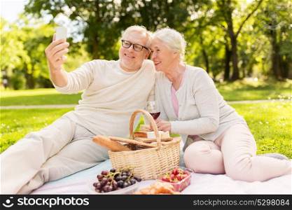 old age, leisure and people concept - happy senior couple with picnic basket and wine taking selfie by smartphone at summer park. senior couple taking selfie at picnic in park