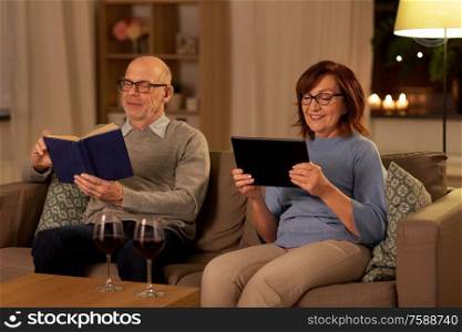 old age, leisure and people concept - happy senior couple with red wine reading book and using tablet pc computer at home in evening. senior couple with book and tablet pc at home