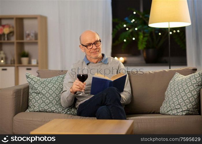 old age, leisure and people concept - happy senior man with glass of red wine reading book at home in evening. happy senior man drinking wine and reading book
