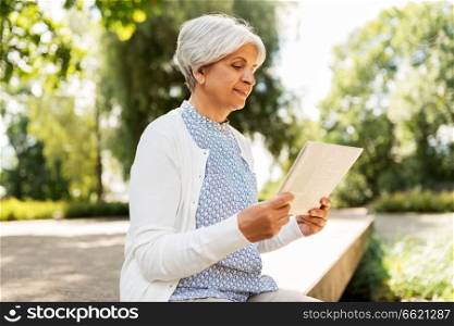 old age, news and mass media concept - senior woman reading newspaper at summer park. senior woman reading newspaper at summer park
