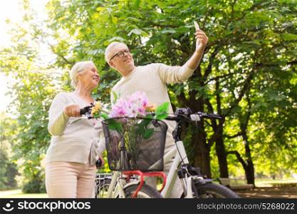 old age, people and lifestyle concept - happy senior couple walking with fixie bicycles taking selfie by smartphone at summer park. senior couple with bicycles taking selfie at park