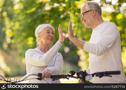 old age, people and lifestyle concept - happy senior couple with fixie bicycles making high five at summer city park. senior couple with bikes making high five at park