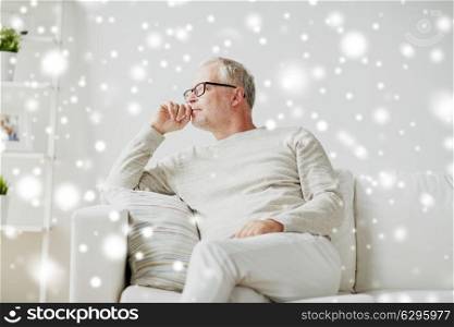 old age, problem and people concept - senior man in glasses thinking and sitting on sofa at home over snow. senior man sitting on sofa at home and thinking