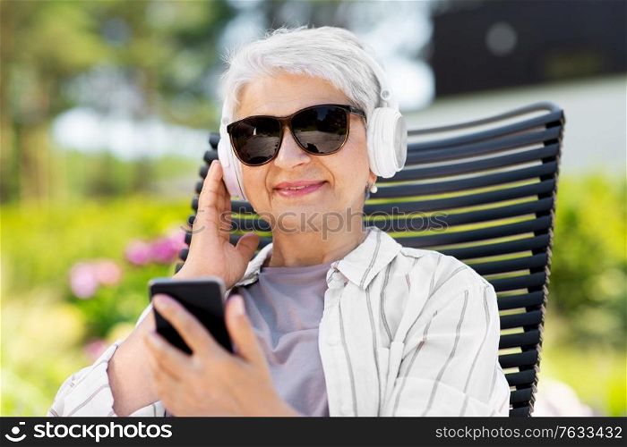 old age, retirement and people concept - happy senior woman in sunglasses with headphones and smartphone listening to music at summer garden. old woman with headphones and smartphone at garden