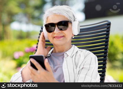 old age, retirement and people concept - happy senior woman in sunglasses with headphones and smartphone listening to music at summer garden. old woman with headphones and smartphone at garden