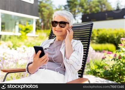 old age, retirement and people concept - happy senior woman in sunglasses with earphones and smartphone listening to music at summer garden. old woman with earphones and smartphone at garden