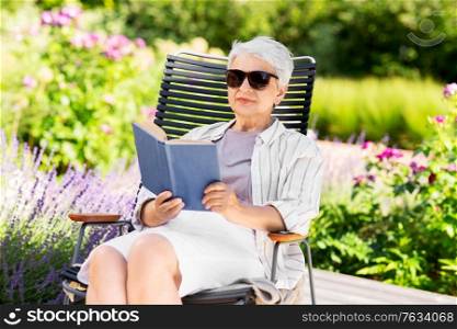 old age, retirement and people concept - happy senior woman reading book sitting in chair at summer garden. happy senior woman reading book at summer garden