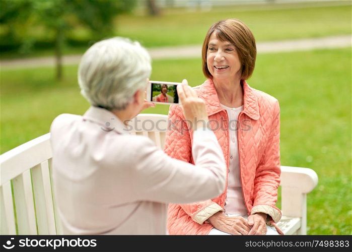 old age, retirement and people concept - senior woman photographing her friend sitting on bench at summer park by smartphone. senior woman photographing her friend at park