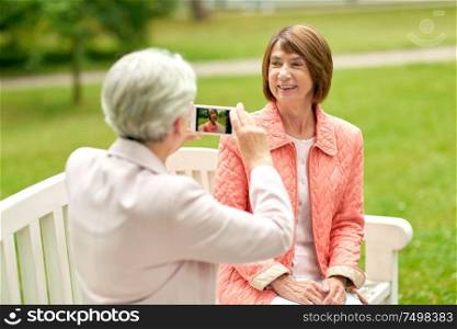 old age, retirement and people concept - senior woman photographing her friend sitting on bench at summer park by smartphone. senior woman photographing her friend at park