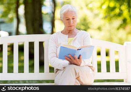 old age, retirement and people concept - senior woman reading book sitting on bench at summer park. senior woman reading book at summer park