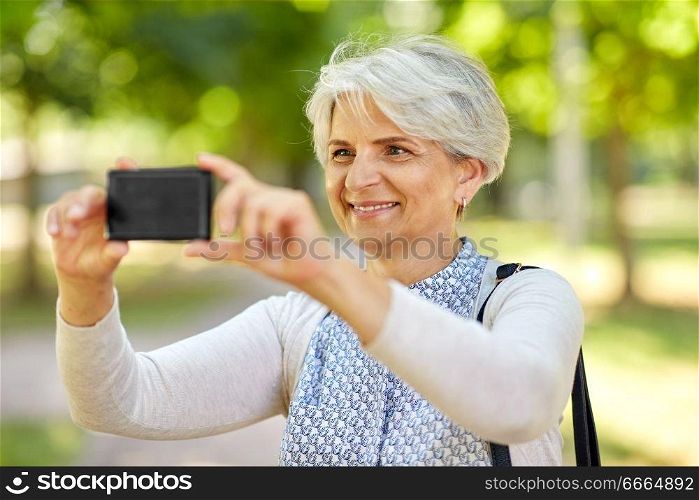 old age, retirement and technology concept - happy senior woman photographing by smartphone summer park. senior woman photographing by cell at summer park