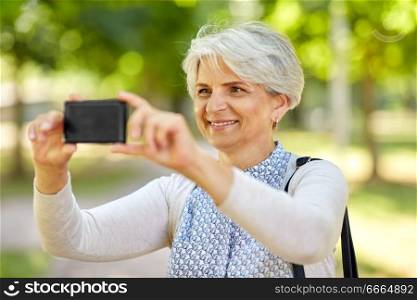 old age, retirement and technology concept - happy senior woman photographing by smartphone summer park. senior woman photographing by cell at summer park
