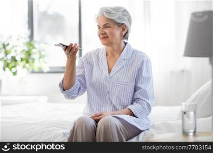 old age, technology and people concept - happy smiling senior woman in pajamas using voice command recorder on smartphone sitting on bed at home bedroom. happy senior woman recording voice on smartphone