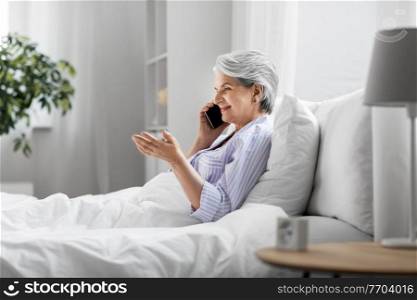 old age, technology and people concept - happy smiling senior woman in pajamas calling on smartphone sitting in bed at home bedroom. senior woman calling on smartphone in bed at home