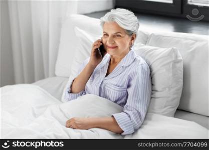 old age, technology and people concept - happy smiling senior woman in pajamas calling on smartphone sitting in bed at home bedroom. senior woman calling on smartphone in bed at home