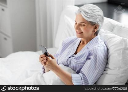 old age, technology and people concept - happy smiling senior woman in pajamas using smartphone sitting in bed at home bedroom. happy senior woman using smartphone in bed at home